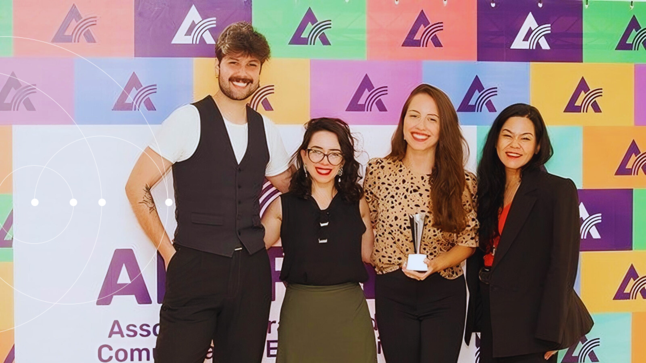Um homem branco e 3 mulheres brancas abraçados enquanto a mulher mais ao centro segura o trófeu da premiação. Eles estão diante de um fundo colorido do evento da Aberje.